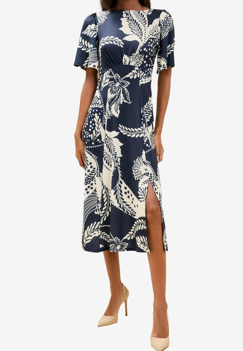 Femme portant une robe midi fleurie bleu marine et blanche, manches courtes et fente latérale haute jusqu'à la cuisse, associée à des talons hauts beiges.