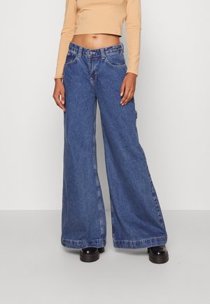 Personne portant un jean bleu taille haute, jambes larges, un top court beige à manches longues et des chaussures noires à plateforme, debout sur un sol uni.