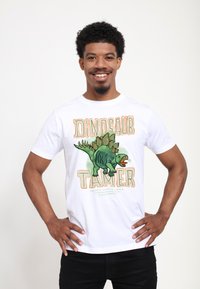 Usměvavý muž s kudrnatými vlasy nosí bílé tričko s obrázkem zeleného stegosaurusa a nápisem "Dinosaur Tamer, Frisco Valley Riverbed California."