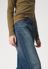 Olijfgroene geribbelde lange mouw top gecombineerd met lichtblauwe denim jeans. De jeans hebben zichtbare naden en een relaxte pasvorm.