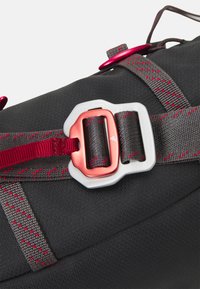 Bolsa de tela negra con cintas grises y una hebilla roja. Presenta costuras rojas y una superficie texturizada, mostrando correas ajustables y herrajes.