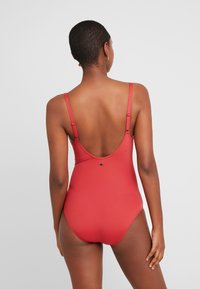 Maillot de bain une pièce rouge avec de fines bretelles réglables, un dos plongeant et une texture lisse, avec un petit logo près de la taille.