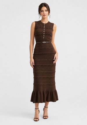 Donna che indossa un vestito midi marrone senza maniche con bottoni decorativi, motivi orizzontali strutturati, vita con cintura e orlo arricciato, con tacchi neri.