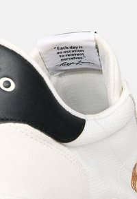 Weißer Sneaker mit strukturierter Oberfläche, schwarzem Lederakzent an der Ferse und einem Stoffetikett mit einem Zitat von Ralph Lauren im Kragen.