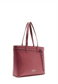 Bolso tote sintético rojo con dos asas largas, detalles de tachuelas en la parte superior y un logo en el frente. Forma rectangular con textura suave.