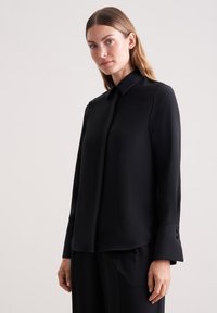 Camicia nera a maniche lunghe con colletto classico, texture liscia e bottoni decorativi sui polsini, abbinata a pantaloni neri.