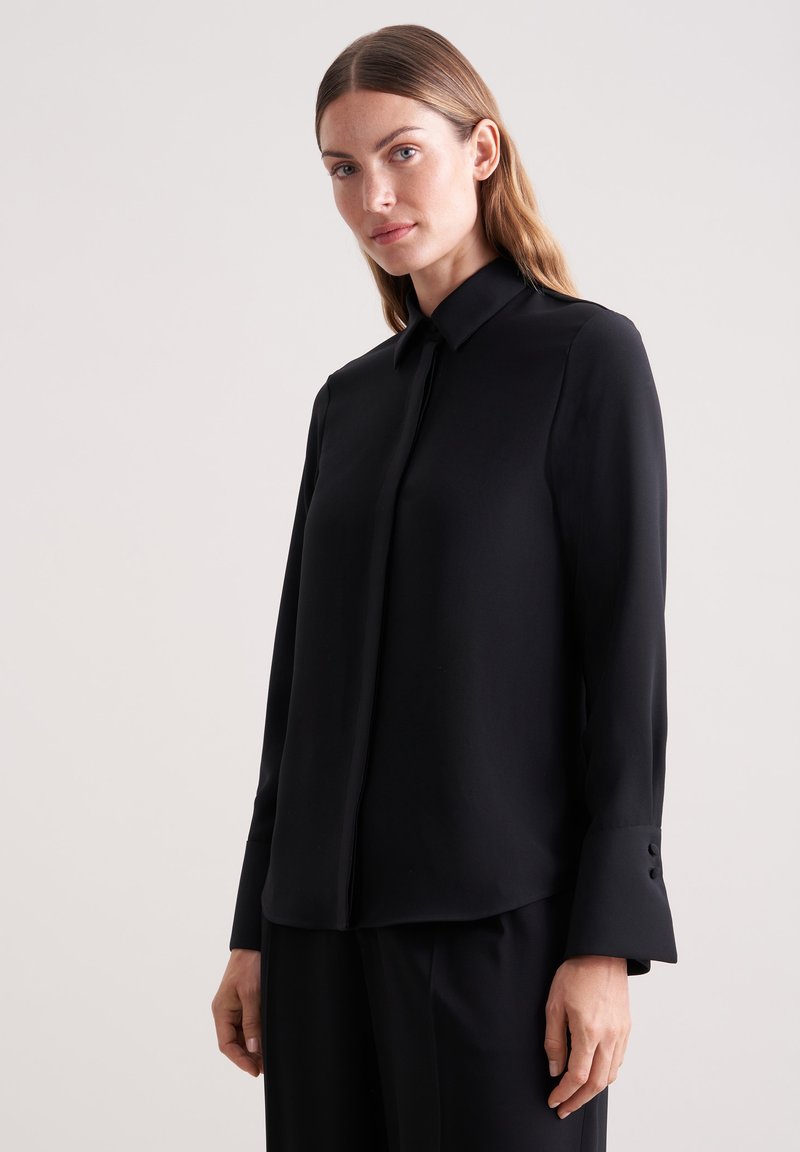 Camicia nera a maniche lunghe con colletto classico, texture liscia e bottoni decorativi sui polsini, abbinata a pantaloni neri.
