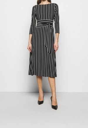 Žena v černých midi šatech s bílými vertikálními pruhy, tříčtvrtečními rukávy, zavazováním v pase a černými lodičkami s špičatou špičkou.