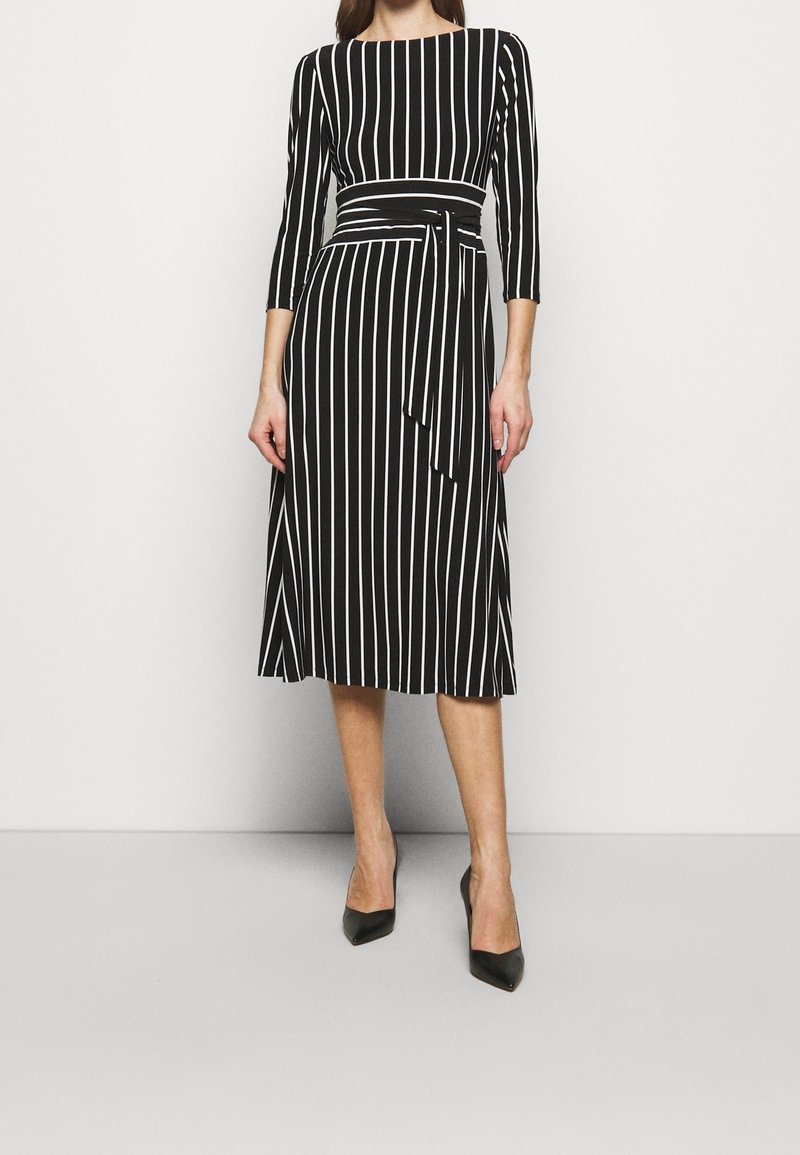 Žena v černých midi šatech s bílými vertikálními pruhy, tříčtvrtečními rukávy, zavazováním v pase a černými lodičkami s špičatou špičkou.