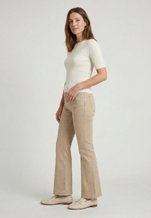 Vrouw staande zijwaarts, gekleed in een witte geribde korte mouw top, beige rechte broek en beige loafers tegen een effen achtergrond.