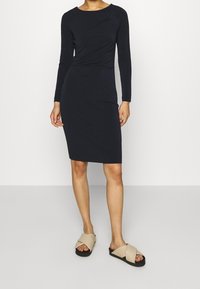 Marinblå stickad långärmad klänning med figursydd siluett och sidovridningsdetalj. Kombineras med beiga sandaler med korsade remmar på en neutral bakgrund.