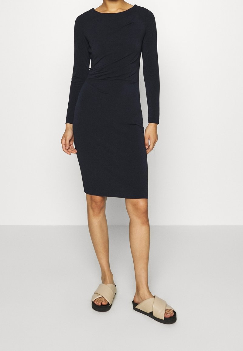 Marinblå stickad långärmad klänning med figursydd siluett och sidovridningsdetalj. Kombineras med beiga sandaler med korsade remmar på en neutral bakgrund.