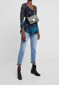Persona que lleva una blusa de estampado azul marino, jeans cortos de color azul claro, botas negras hasta el tobillo y un bolso cruzado verde oliva con un cierre cuadrado.