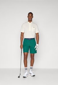 Polo shirt crem cu un buzunar mic și un logo, pantaloni șort verzi, curea bleumarin, șosete în dungi, încălțăminte de golf albă, ținând un club de golf.