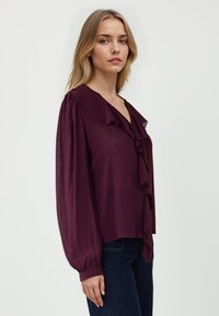 Blusa bordeaux con fronte arricciato, scollatura a V profonda e maniche lunghe e voluminose. Realizzata in un tessuto leggero e fluente.