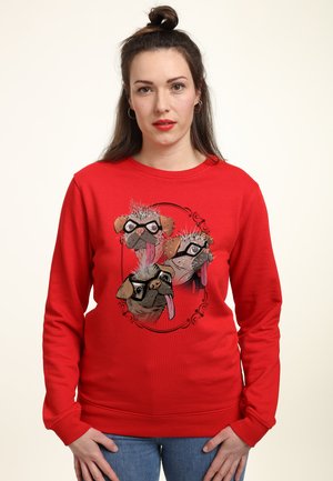 Kvinde med mørkt hår i en topknude iført en rød sweatshirt med tre tegneserie-mops ansigter med briller trykt på fronten.
