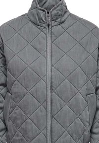 Veste grise matelassée avec un col haut et une fermeture éclair à l'avant. Le matériau est doux avec un motif en diamant et une texture légèrement rembourrée.