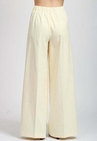 Persona in pantaloni larghi a vita alta color crema, in piedi con la schiena rivolta verso la macchina fotografica su uno sfondo neutro.