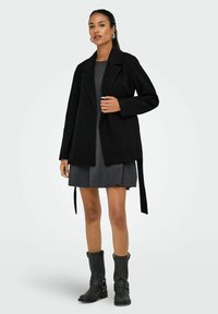 Manteau noir avec revers, en mélange de laine, associé à une robe grise. Le mannequin porte des bottes noires jusqu'aux genoux avec des boucles.