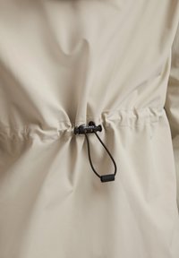 Beige jasje detail met een aangetrokken taille, uitgerust met een zwart trekkoordmechanisme dat een gelabelde plastic toggles heeft en een gladde textuur.