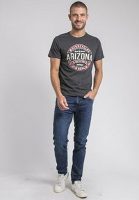 T-shirt en coton gris charbon avec un graphique circulaire rouge et crème portant "Arizona" et des motifs de moto, associé à un jean bleu foncé et des baskets blanches.