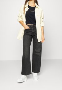 Svarta vida jeans kombinerade med en svart kortad topp med guldtext och en öppen beige jacka. Vita sneakers fulländar outfiten.