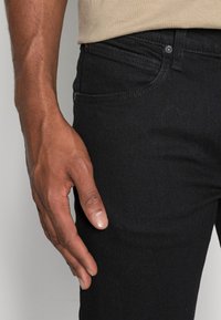 Mustat farkut, joissa on sileä pinta, slim-fit-malli ja näkyvät ompeluyksityiskohdat. Yksi käsi lepää reisi alueella.