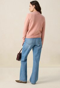 Pull en maille rose avec un col, associé à un jean évasé bleu clair. Portant un sac marron et des chaussures à motifs. Fond neutre.