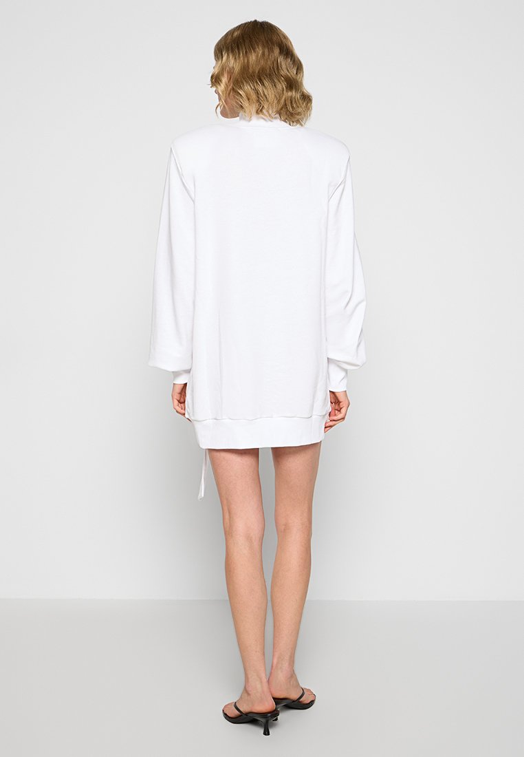 Osoba stojąca plecami, ubrana w długą, białą, oversize'ową sukienkę swetrową oraz czarne sandały na obcasie z odkrytą palcą, na jednolitym tle.