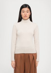 Lyse beige høyhalset genser laget av mykt strikket materiale, med lange ermer og ribbestrikket kant, sammen med brune plissert bukser.