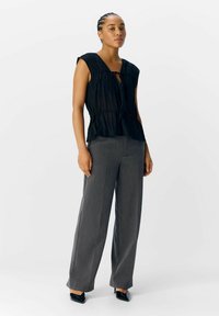 Top negro con pliegues, escote en V y detalle de lazada, combinado con pantalones de pierna ancha grises y tacones negros puntiagudos. Textura suave, diseño en capas.