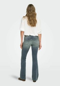 Palaidiniai džinsai su išplatėjusia apačia, turintys gradientinį mėlyną skalbimą, su užpakalinėmis kišenėmis ir liekna forma ant klubų. Derinama su balta bluzo.