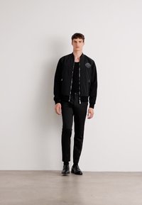 Black bomber jacket with a textured finish, features a logo on the chest, paired with fitted black pants and black dress shoes.