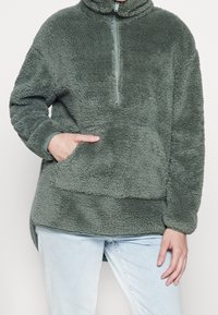 Zielony polarowy sweter z pół-zamkiem, stójką i przednią kieszenią. Miękka tekstura, luźny krój i boczne rozcięcia u dołu.