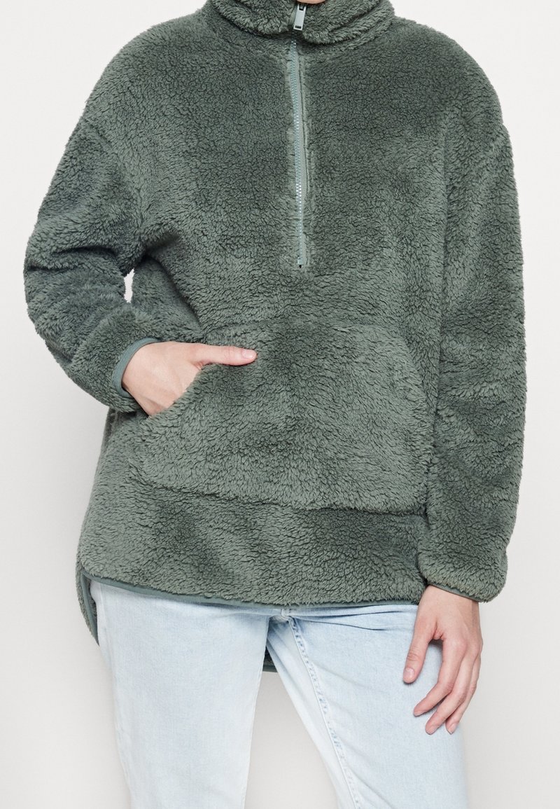 Zielony polarowy sweter z pół-zamkiem, stójką i przednią kieszenią. Miękka tekstura, luźny krój i boczne rozcięcia u dołu.