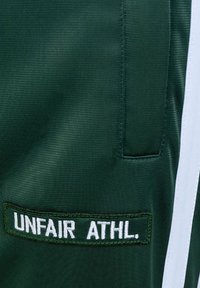 Tissu vert foncé avec un patch cousu portant l'inscription "UNFAIR ATHL." et une bande blanche verticale sur le côté droit.