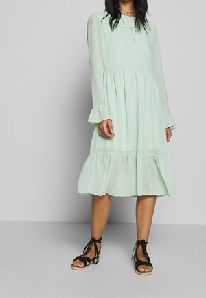 Woman wearing a light green long-sleeve midi dress with ruffled hem and black lace-up sandals, standing against a gray background.