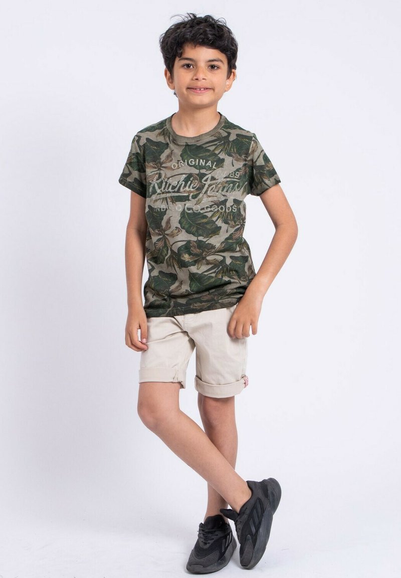 T-shirt camouflage avec un motif de feuilles vertes et brunes, à manches courtes, associé à un short beige retroussé et des baskets noires.