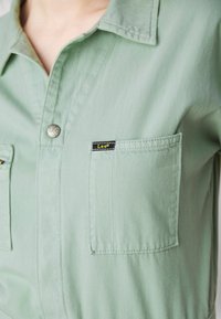 Chemise boutonnée vert clair avec poches poitrine, dont une étiquetée avec une petite marque Lee en noir et jaune.