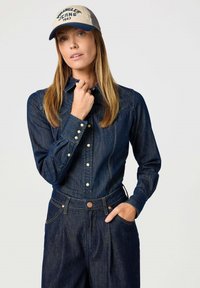 Camisa de mezclilla oscura con cuello, botones de presión de perla y mangas largas. Combinada con pantalones de mezclilla oscura de cintura alta y una gorra beige.
