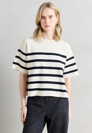 Joven con cabello rubio que lleva una camiseta blanca de manga corta con rayas horizontales negras y pantalones oscuros, de pie contra un fondo claro.