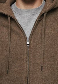 Sweat à capuche marron zippé en tissu texturé, avec deux cordons de serrage et une fermeture éclair argentée. Un sweat-shirt gris est superposé en dessous.