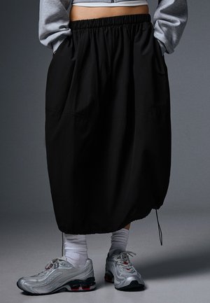 Personne portant une longue jupe noire à taille élastique, un sweat-shirt court gris, des chaussettes blanches et des chaussures de sport argentées, debout sur un sol gris.