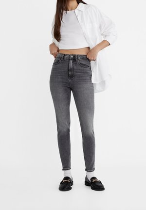 Femme portant un jean skinny gris taille haute, un débardeur blanc court, une chemise blanche déboutonnée, des chaussettes blanches et des mocassins noirs avec un détail doré.
