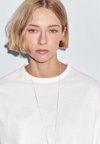 Young person with short blonde hair, wearing a white shirt and thin silver necklace, looking straight ahead against a plain light background.