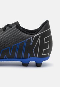Zapatilla de fútbol negra con material texturizado, presenta un logo de Nike azul y acentos grises, forma redondeada y una suela azul con tacos negros.