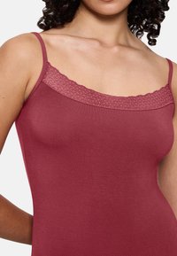 Kobieta w bezrękawniku w kolorze przybrudzonego różu z koronkowym wykończeniem dekoltu, odsłaniająca ramiona i górną część klatki piersiowej na jednolitym tle.