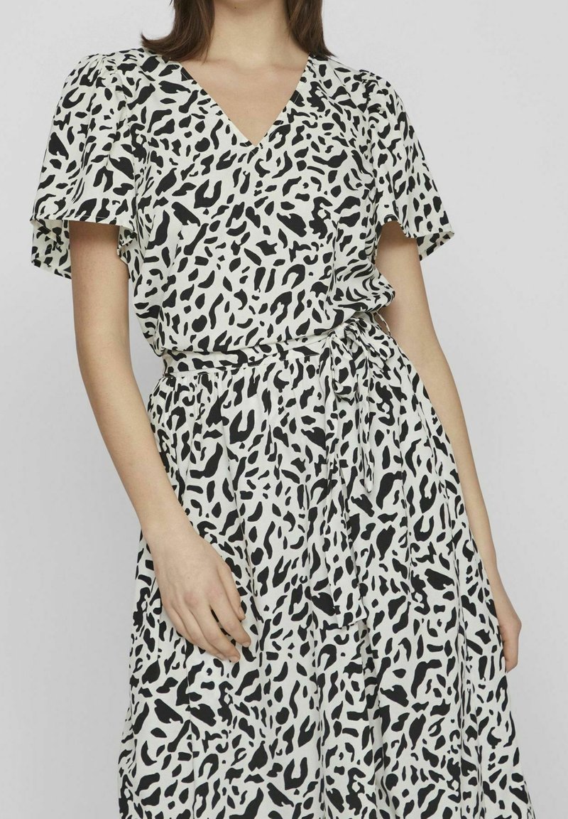 Femme portant une robe à imprimé léopard noir et blanc avec manches courtes flottantes et ceinture nouée à la taille, sur fond uni.