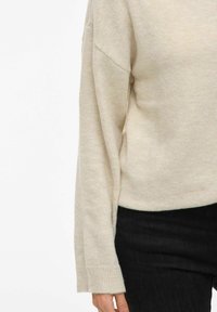 Beige pulover s dolgimi rokavi in rebrastimi manšetami, oblečen z črnimi hlačami, prikazuje delni pogled na trup in roko.