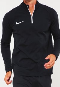 Pull-over Nike noir à manches longues avec un col zippé sur un quart, arborant un logo blanc sur la poitrine et une coupe lisse et ajustée.